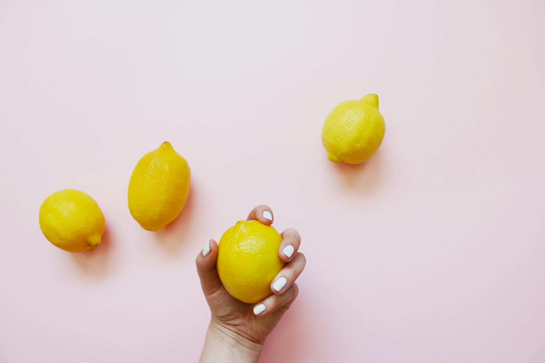A hand with white nails holding a lemon on a soft pink background, symbolizing fresh, clean self-care.