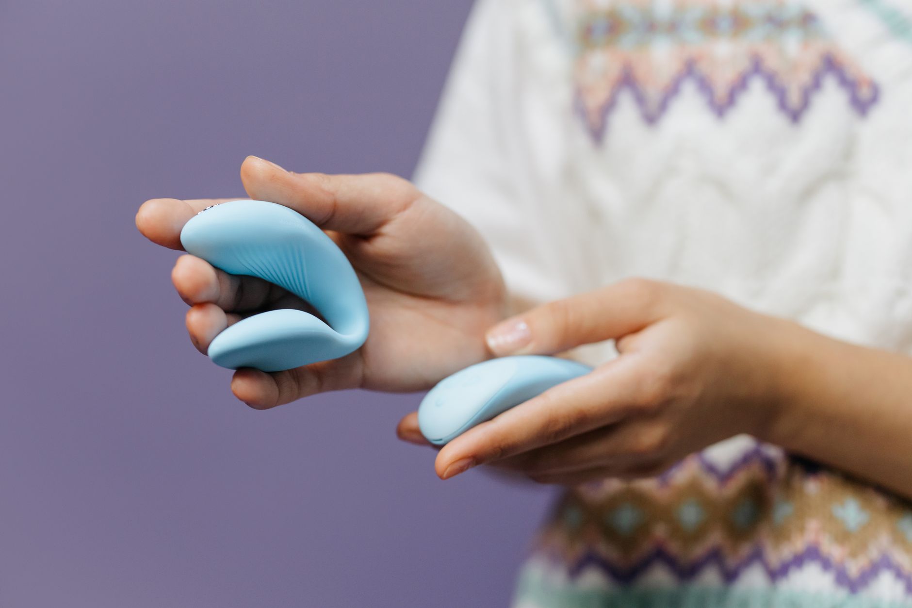 Close-up of hands holding a sleek blue clitoral vibrator against a purple background, representing shared exploration